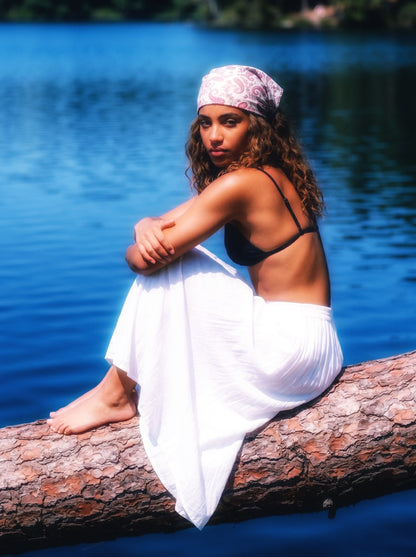 Femme assise sur un tronc au bord d’un lac, portant un bandana clair à motif paisley, scène naturelle et lumineuse.